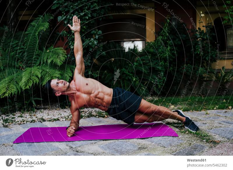Shirtless male athlete practicing plank position on mat against plants in yard color image colour image Spain outdoors location shots outdoor shot outdoor shots