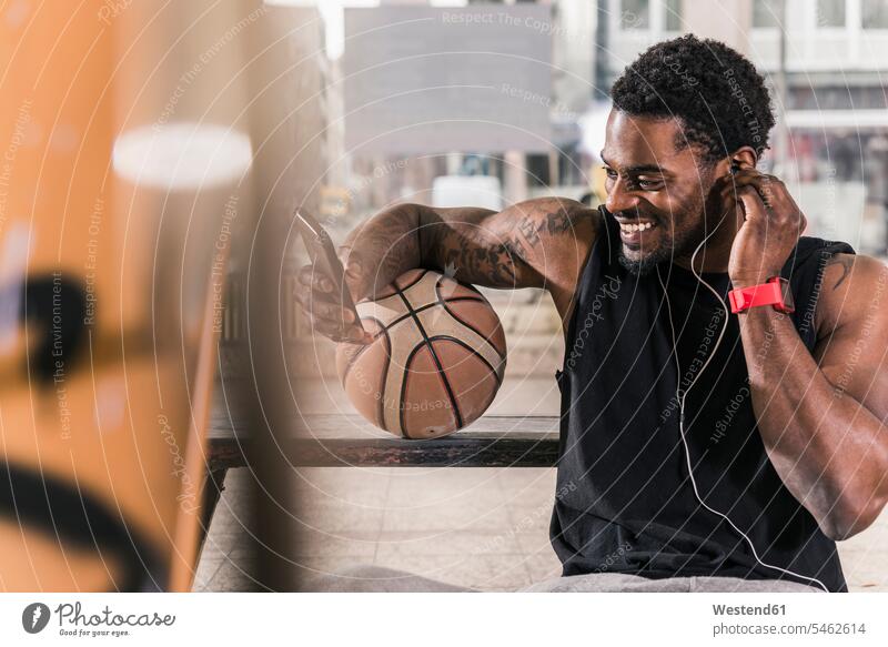 Smiling man with tattoos and basketball using smartphone and earphones ear phone ear phones men males Smartphone iPhone Smartphones smiling smile basketballs