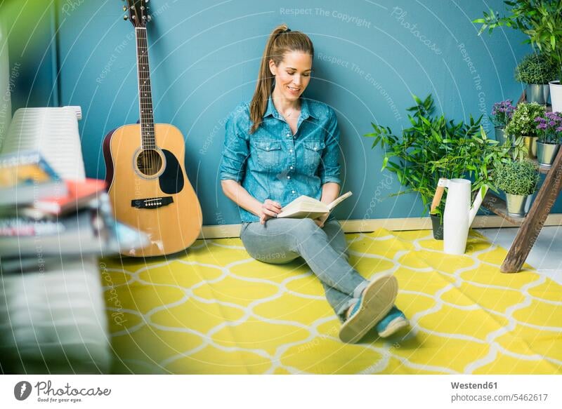 Woman relaxing in her home with potted plants, reading a book relaxation relaxed at home woman females women sitting Seated guitar guitars books Adults