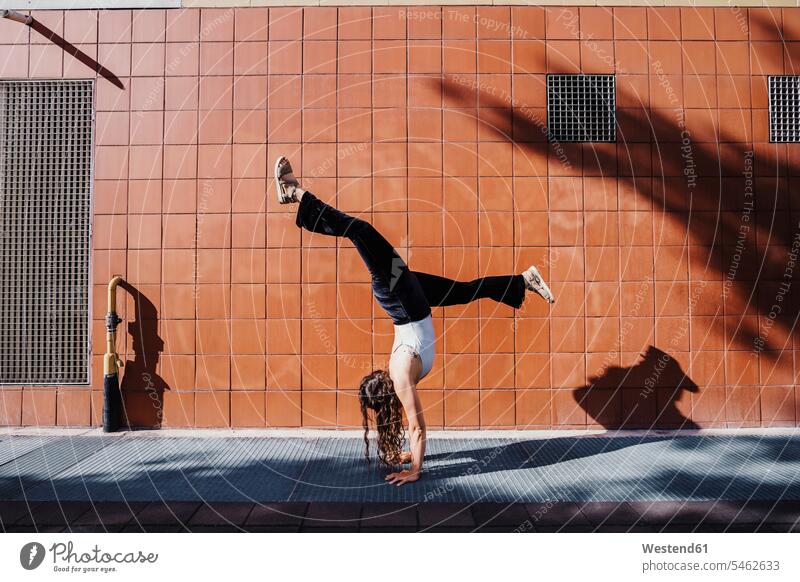 Young woman performing handstand on sidewalk against tiled wall in city during sunny day color image colour image outdoors location shots outdoor shot
