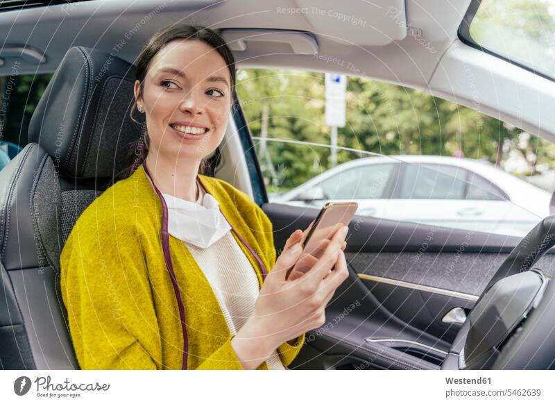 Portrait of smiling woman in car with protective mask and smartphone coat coats jackets motor vehicles road vehicle road vehicles Auto automobile Automobiles