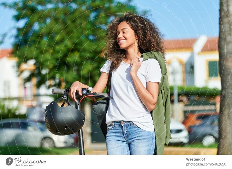 Happy woman carrying jacket while standing with electric push scooter during sunny day color image colour image outdoors location shots outdoor shot