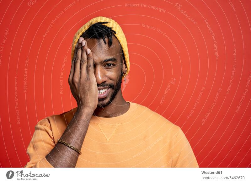Portrait of stylish young man covering one eye in front of orange wall heads faces human face human faces jumper sweater Sweaters smile delight enjoyment