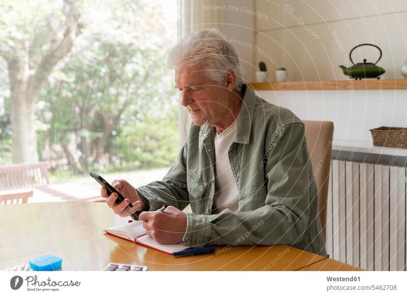 Senior diabetic man using mobile phone while writing in diary at home color image colour image indoors indoor shot indoor shots interior interior view Interiors