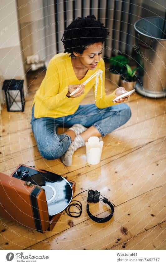 Young woman at home sitting on with record player, checking smarphone while eating noodles young women young woman turntable message Seated Smartphone iPhone