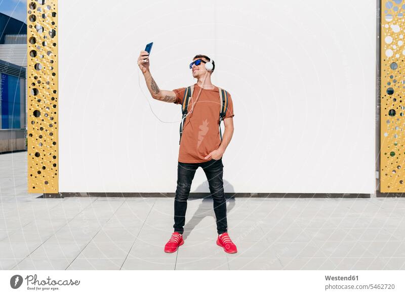 Smiling man wearing headphones taking selfie through mobile phone while standing with hands in pockets against wall color image colour image outdoors