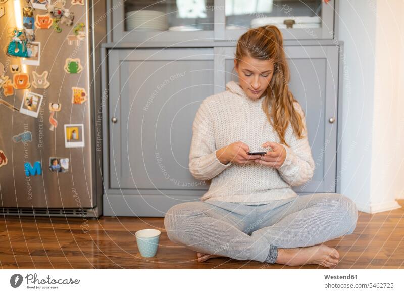 Young woman sitting on the floor in kitchen at home using cell phone human human being human beings humans person persons celibate celibates singles