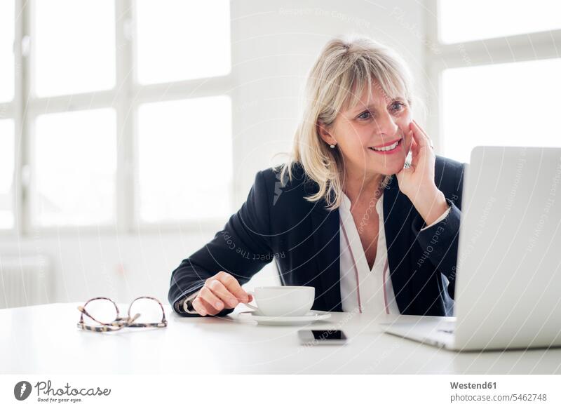 Smiling mature businesswoman with cup of coffee working on laptop at desk in the office Laptop Computers laptops notebook Coffee Cup Coffee Cups desks