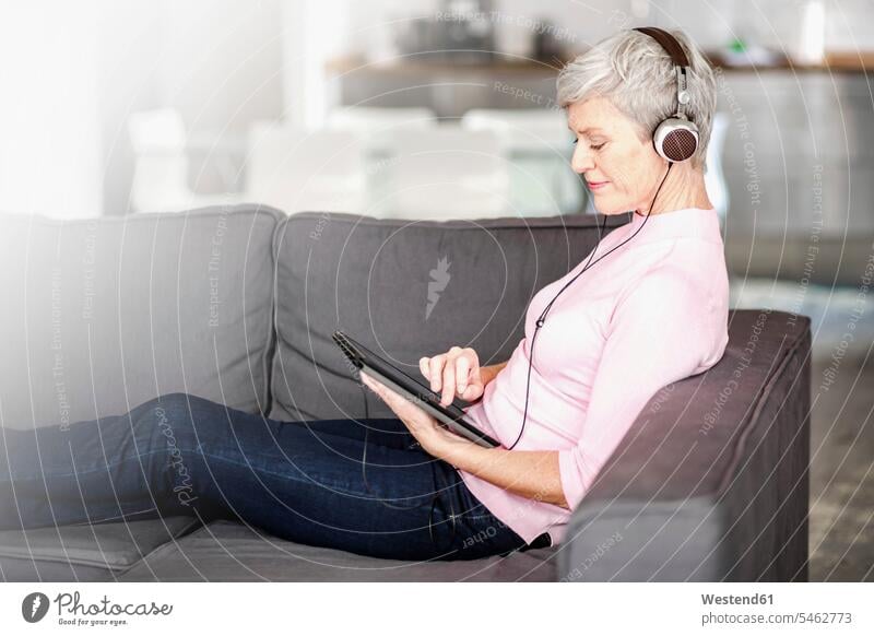 Mature woman sitting on couch using digital tablet while listening music with headphones Seated use females women hearing digitizer Tablet Computer Tablet PC
