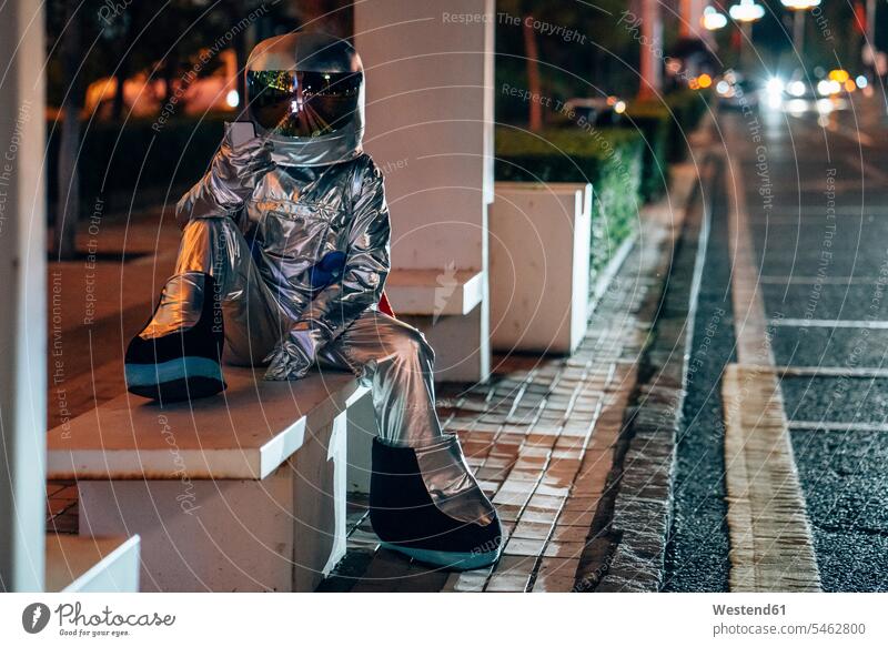 Spaceman sitting on bench at a bus stop at night holding cell phone astronaut astronauts busstops road streets roads spaceman spacemen city town cities towns
