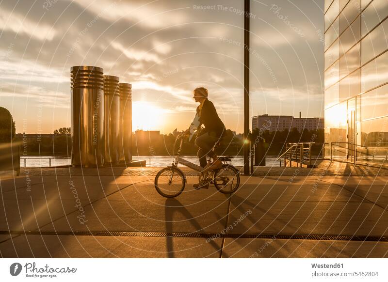 Senior woman riding city bike at the riverside at sunset sunsets sundown riverbank bicycle bikes bicycles atmosphere atmospheric mood moody Atmospheric Mood