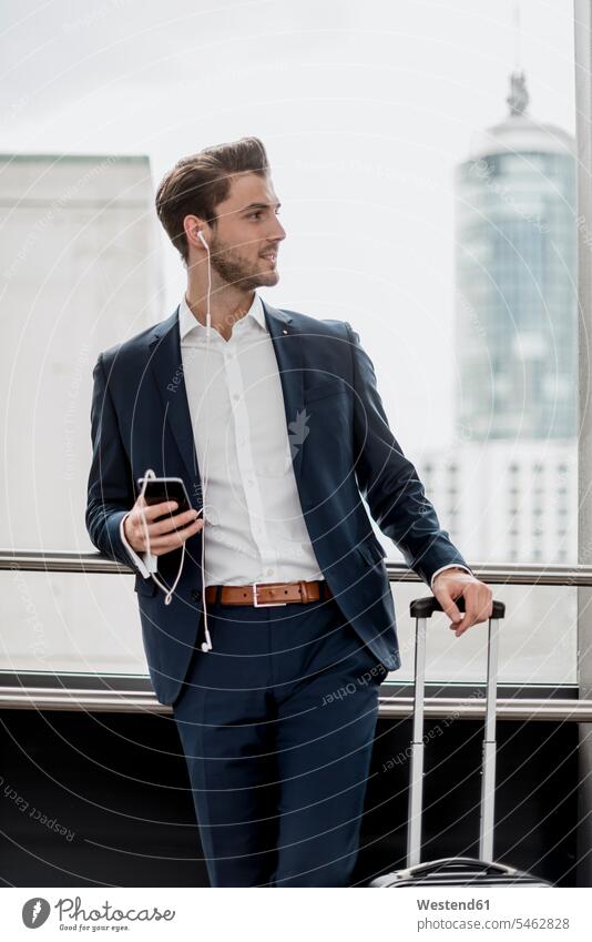 Young businessman at the window with cell phone, earbuds and rolling suitcase Businessman Business man Businessmen Business men rolling suitcases windows