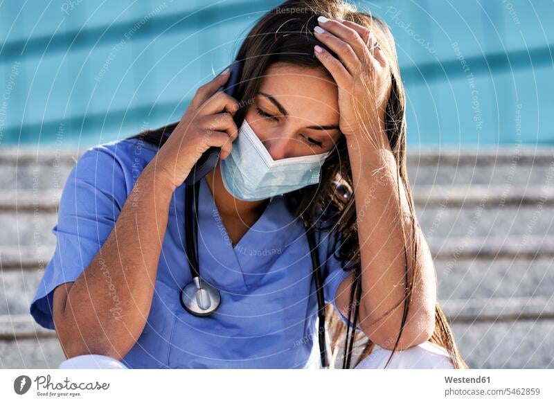 Young doctor wearing face mask talking on mobile phone while sitting on staircase against hospital color image colour image outdoors location shots outdoor shot