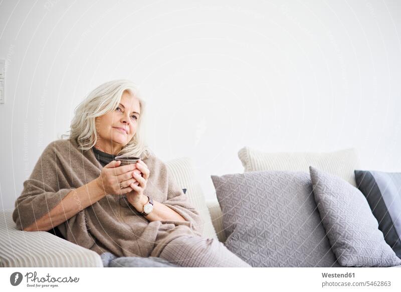Thoughtful retired woman holding coffee cup while sitting on sofa against white wall at apartment color image colour image Denmark Scandinavia