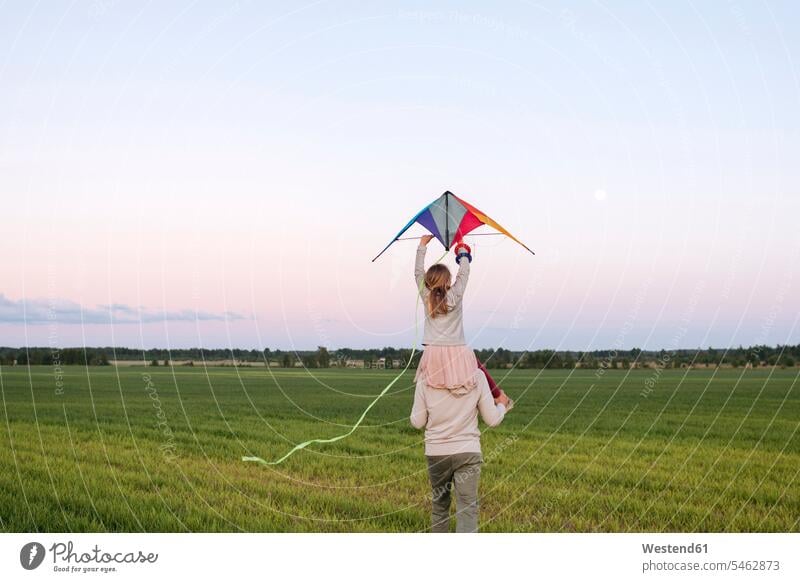 Man carrying daughter holding kite on shoulders while walking on grassy landscape at sunset color image colour image leisure activity leisure activities