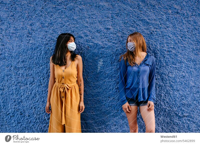 Female friends wearing masks looking at each other while maintaining distance against blue wall in city color image colour image Spain leisure activity