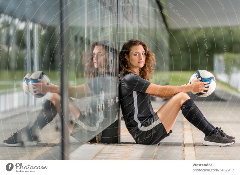 Female soccer player holding ball while sitting on footpath by glass wall color image colour image Germany outdoors location shots outdoor shot outdoor shots