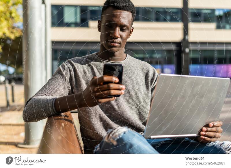 Young man sitting on a bench in the city, using laptop and smartphone Laptop Computers laptops notebook Seated benches Smartphone iPhone Smartphones