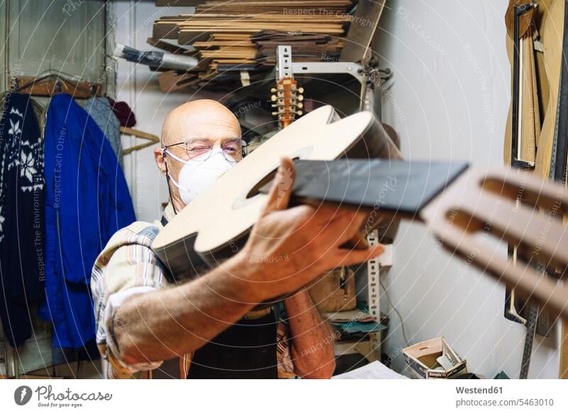 Craftsman with face mask examining guitar while standing at workshop color image colour image indoors indoor shot indoor shots interior interior view Interiors
