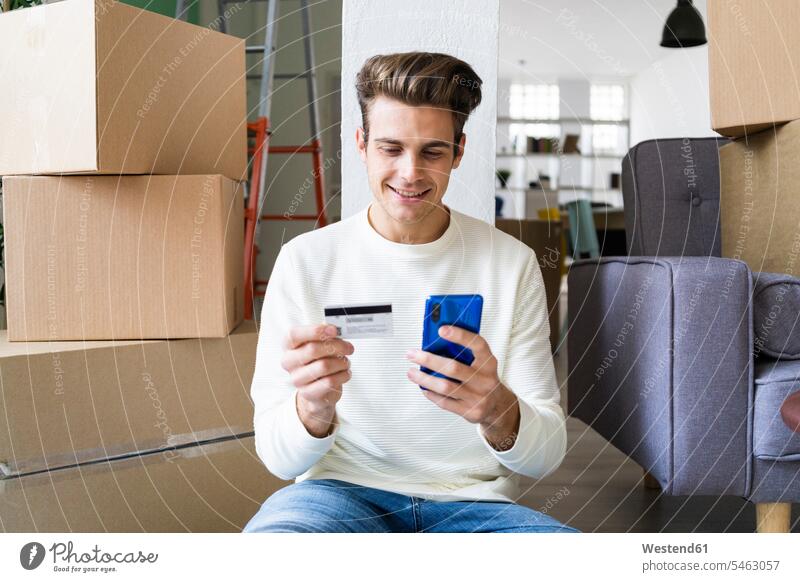Smiling young man paying through mobile phone by credit card while moving in new house color image colour image indoors indoor shot indoor shots interior