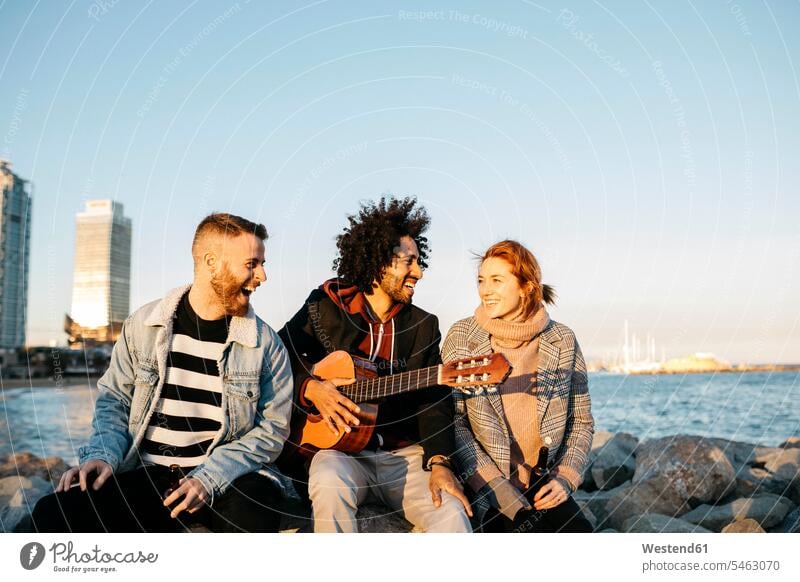 Three happy friends with guitar sitting outdoors at the coast sunset happiness Seated coastline shoreline sunsets sundown guitars friendship atmosphere