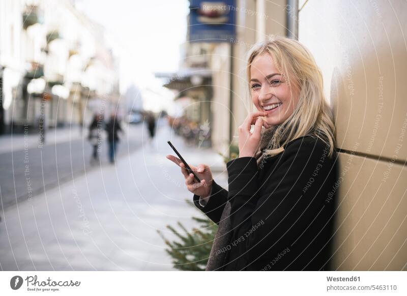 Portrait of smiling young woman with cell phone in the city blond blond hair blonde hair Coat Coats wireless Wireless Connection Wireless Technology