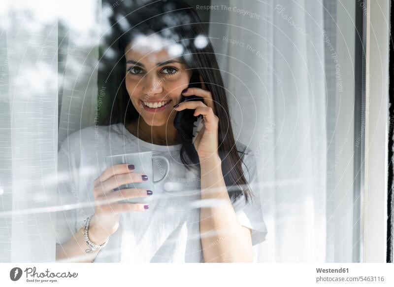 Portrait of smiling young woman on cell phone behind windowpane human human being human beings humans person persons celibate celibates singles solitary people