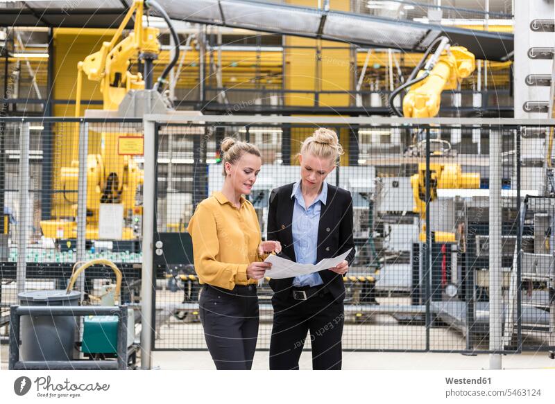 Two women discussing plan in factory shop floor with industrial robot woman females plans industrial hall factory hall industrial buildings factories discussion