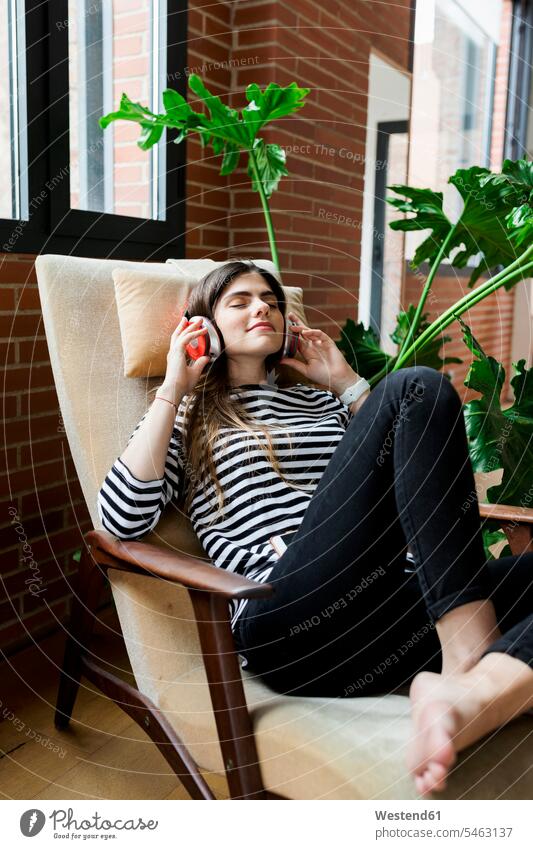 Young woman sitting in armchair at home listening to music with headphones headset hearing females women Arm Chairs armchairs Seated Adults grown-ups grownups