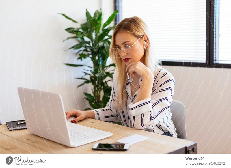 Blond office worker sitting at desk, using laptop Seated desks blond blond hair blonde hair using a laptop Using Laptops working At Work offices office room