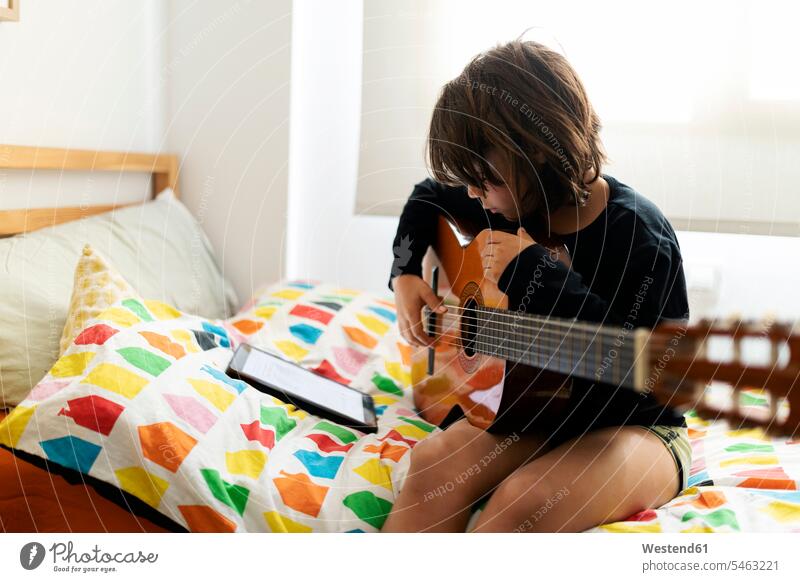 Boy sitting on bed using digital tablet for playing song on guitar Instrument Instruments musical instruments stringed instruments guitars acoustic guitars