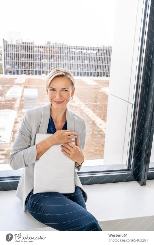 Blond businesswoman with laptop, sitting on windowsill in office building human human being human beings humans person persons caucasian appearance