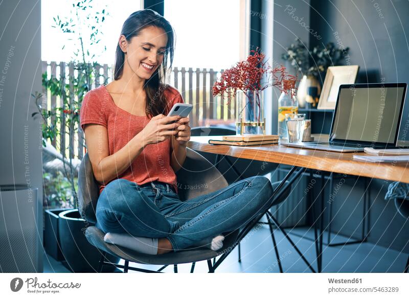 Female entrepreneur using smart phone while sitting on chair by desk in home office color image colour image casual clothing casual wear leisure wear