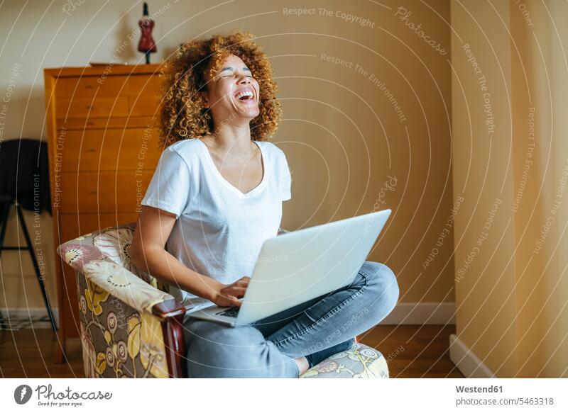 Happy young woman with curly hair sitting in armchair at home using laptop Seated females women Laptop Computers laptops notebook Arm Chairs armchairs curls