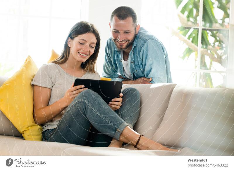 Happy couple at home in modern living room on couch looking at tablet together windows cushions couches settee settees sofa sofas hold smile Seated sit relax