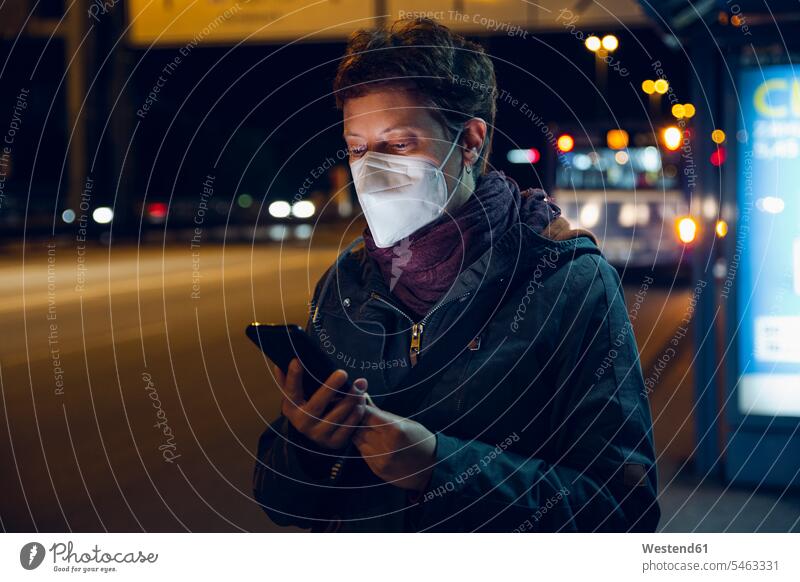Woman wearing protective face mask using smart phone while standing at bus stop during COVID-19 color image colour image outdoors location shots outdoor shot