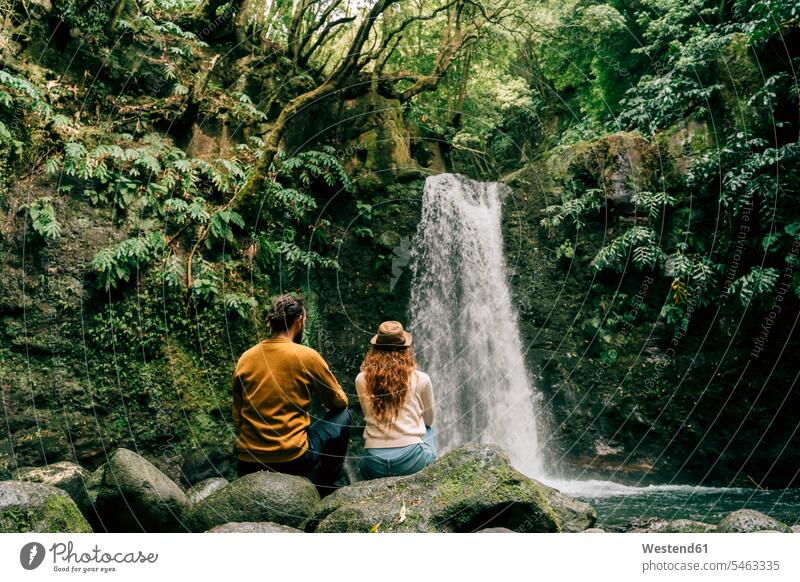 Rear view of couple sitting at a waterfall on Sao Miguel Island, Azores, Portugal touristic tourists Seated enjoy enjoyment indulgence indulging savoring