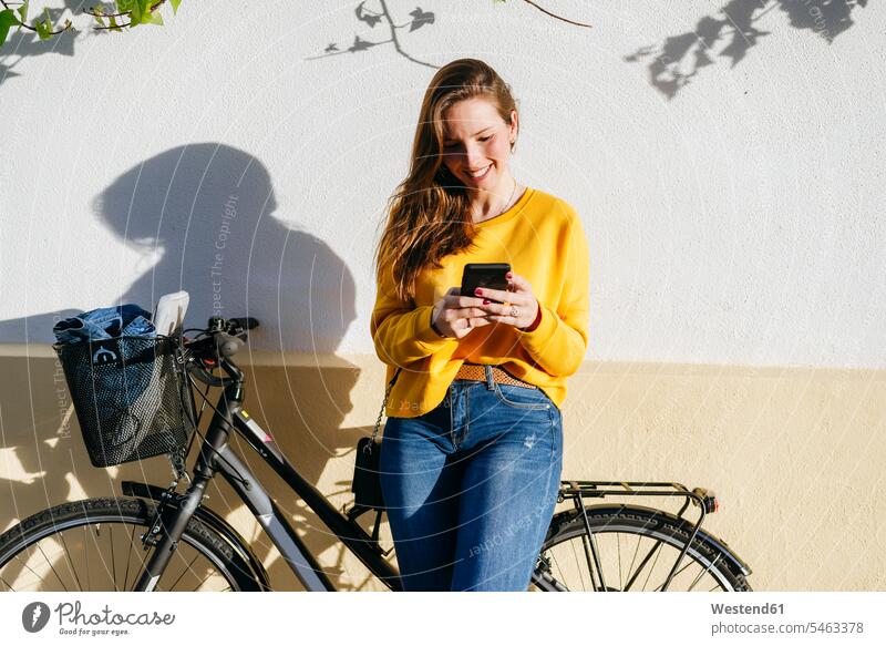 Smiling young woman with bicycle using cell phone at a wall females women walls bikes bicycles mobile phone mobiles mobile phones Cellphone cell phones Adults