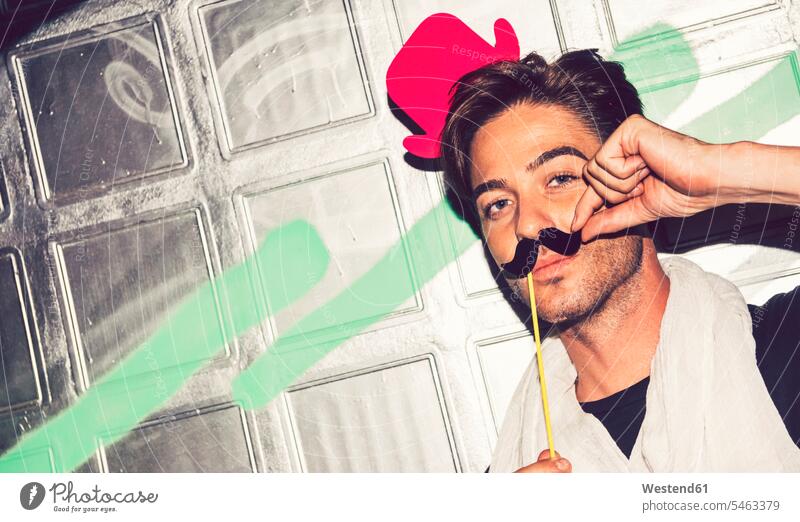 Close-up of handsome man wearing fake hat with mustache against graffiti wall color image colour image Spain leisure activity leisure activities free time