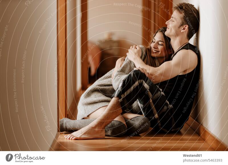 Happy young couple sitting on the floor at home jumper sweater Sweaters cuddle snuggle snuggling smile Seated embrace Embracement hug hugging relax relaxing