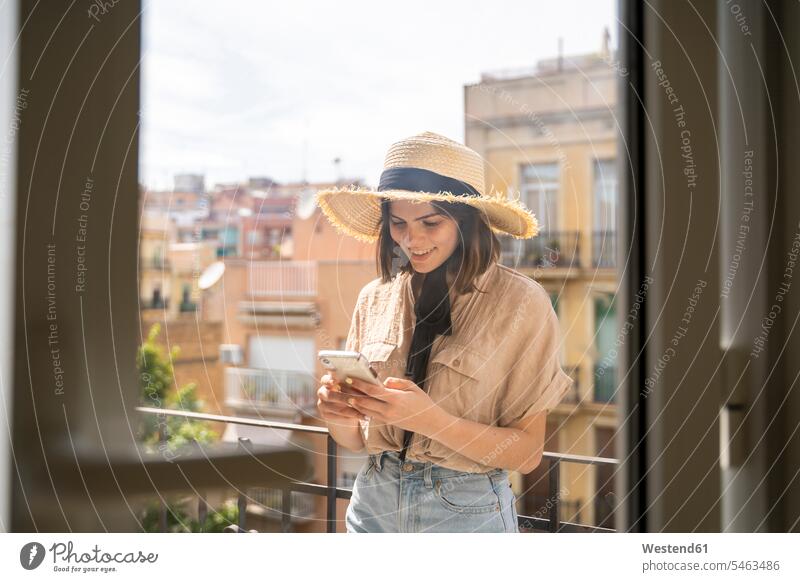 Smiling young woman on balcony in the city wearing straw hat using cell phone shirts telecommunication phones telephone telephones cell phones Cellphone mobile