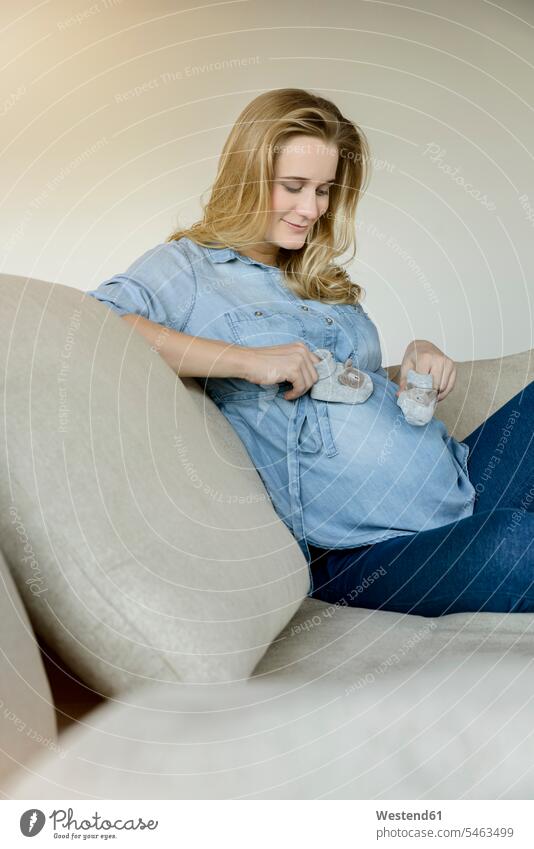 Smiling pregnant woman sitting on couch holding baby shoes Seated females women Pregnant Woman settee sofa sofas couches settees Baby Shoe baby booties home