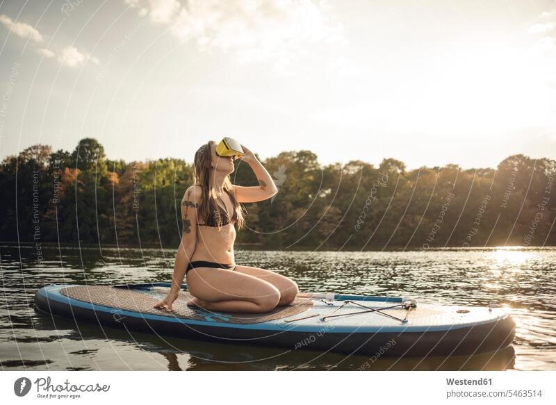 Young woman with tatto kneeling on paddleboard, using VR goggles swim wear bikinis aquatics Water Sport surf board surf boards surfboards relax relaxing