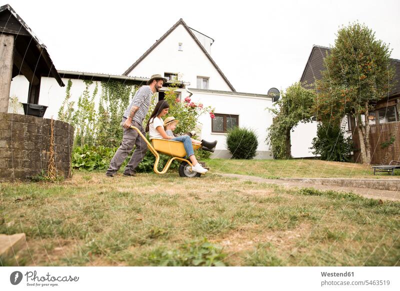 Playful man pushing wife and son sitting in wheelbarrow in garden family families Seated hand barrow pushcart wheelbarrows hand barrows wheel barrow gardens