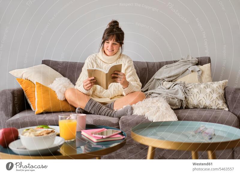 Smiling woman in sweater reading book while sitting on sofa at home color image colour image Spain indoors indoor shot indoor shots interior interior view
