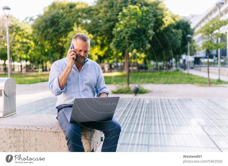 Businessman using laptop while talking on mobile phone at office park business people businesspeople Business Professional Business Professionals Businessperson