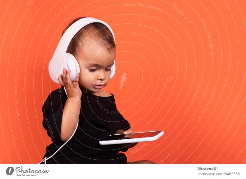 Cute baby girl wearing headphones using mobile phone while sitting against orange background color image colour image studio shot studio photograph