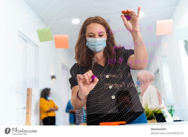 Young woman writing on screen partition while standing with coworker in background at office color image colour image indoors indoor shot indoor shots interior