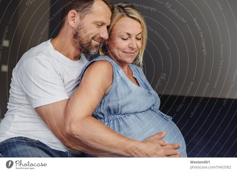 Mature man and his pregnant mature wife sitting on bed touching her belly Pregnant Woman Seated beds bellies abdomen human abdomen upper body