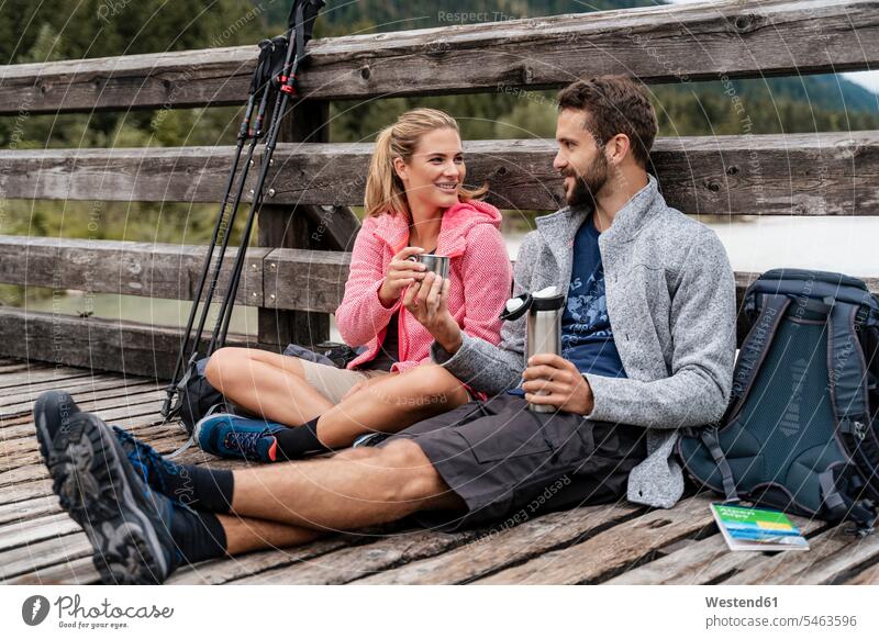 Young couple having a break on a wooden bridge during a hiking trip, Vorderriss, Bavaria, Germany touristic tourists cup mugs back-pack back-packs backpacks